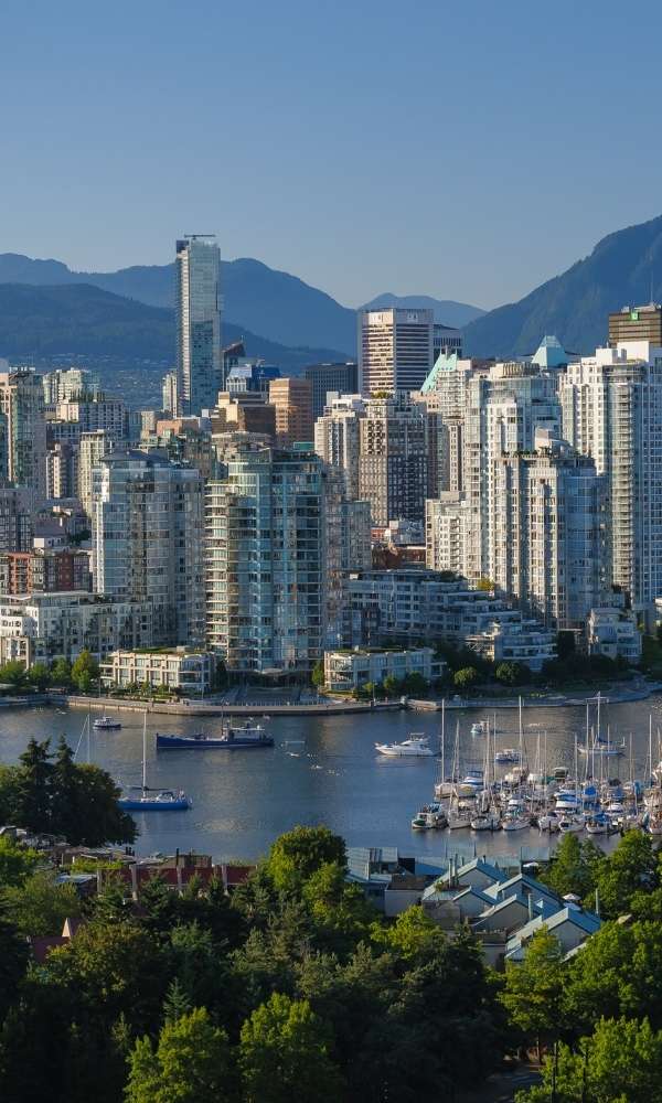 A scenic photo of Vancouver BC’s skyline with mountains, water, and modern high-rise buildings under a blue sky.