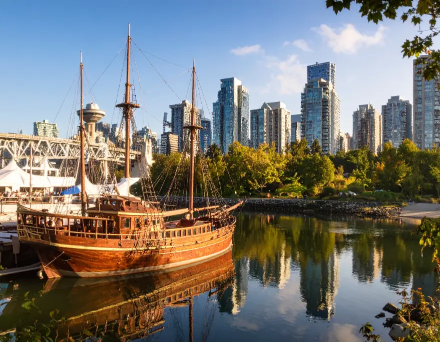 A vintage wooden ship docked near Vancouver BC’s skyline, surrounded by trees and water with mountains in the distance.