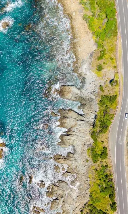 Winding coastal road hugging a rocky shoreline with crashing waves and vibrant blue ocean under clear skies.